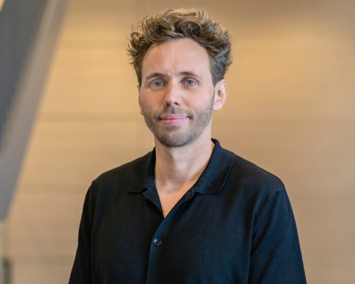 Mann mit lockigem Haar in schwarzem Shirt, steht vor einem hellen Hintergrund.