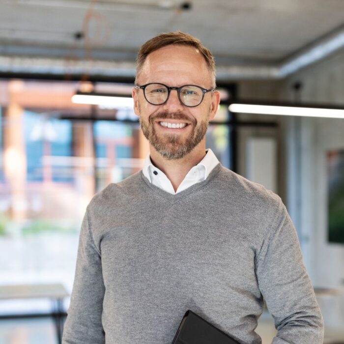 Mann mit Brille und grauem Pullover steht in einem modernen Büro mit großen Fenstern und einem Tablet in der Hand.