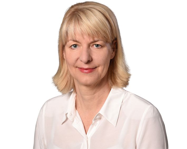 Porträt einer Frau mit blonden Haaren, die in einem weißen Hemd vor einem weißen Hintergrund lächelt.