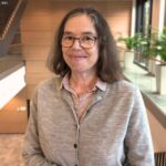 Frau mit Brille und braunen Haaren, trägt einen grauen Pullover, steht in einem modernen Innenraum mit Pflanzen.