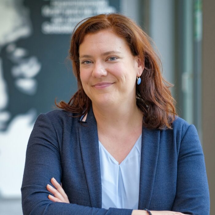 Johanna Wenckebach steht im Foyer der University of Labour. Sie schaut selbstbewusst in die Kamera. Im Hintergrund ist das Portrait von Hugo Sinzheimer an der Wand zu erkennen.