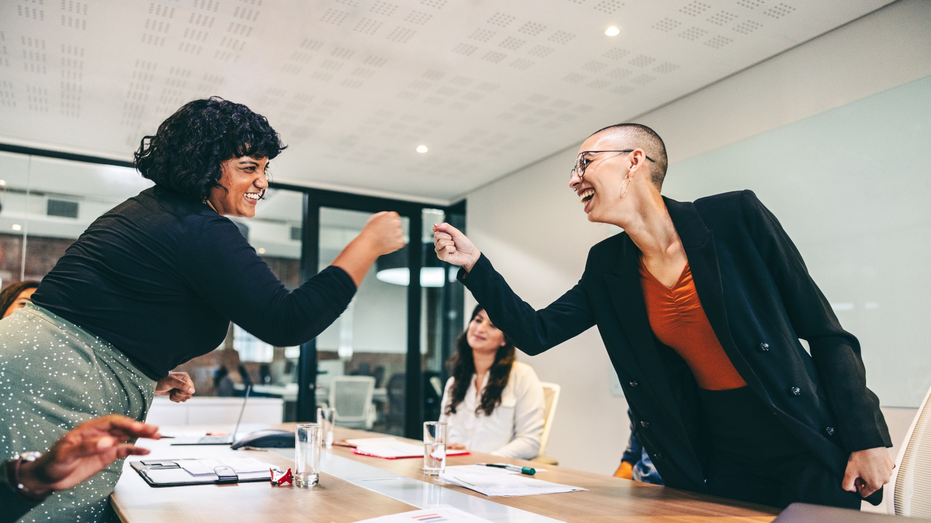 Zwei Personen fausten einander zu. Im Mittelpunkt des Bildes sehen wir, wie die beiden Fäuste sich fast berühren. Links und rechts sehen wir jeweils die beiden Personen. Im Umfeld sehen wir eine Meeting-Situation in einem Büro.
