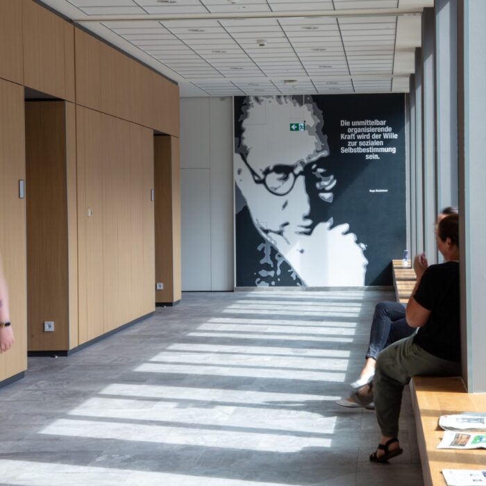 Person geht durch einen hellen Flur in der University of Labour mit Holzpaneelen, an der Wand ein großes Porträt und Zitat von Hugo Sinzheimer.