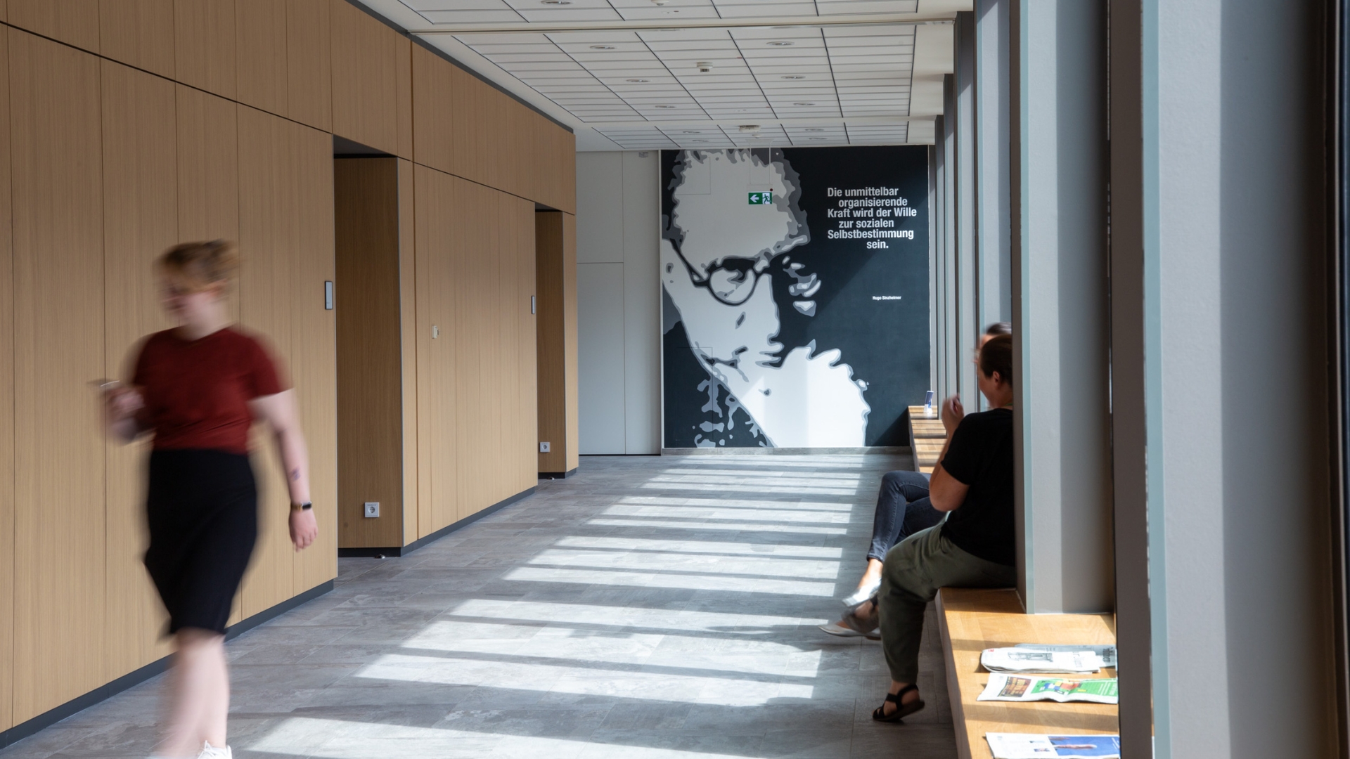Person geht durch einen hellen Flur in der University of Labour mit Holzpaneelen, an der Wand ein großes Porträt und Zitat von Hugo Sinzheimer.
