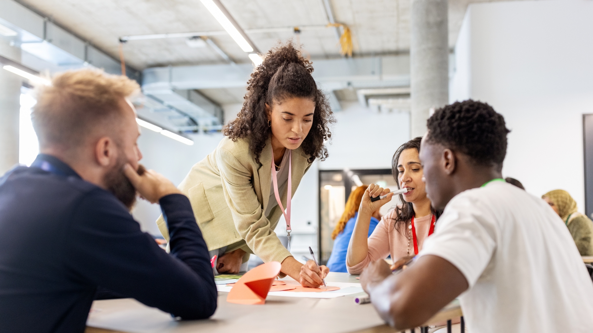 Vier Personen arbeiten zusammen an einem Tisch. Alle überlegen gemeinsam. Drei Personen sitzen, eine Frau in der Mitte des Bildes beugt sich stehend leicht über den Tisch und zeigt etwas.