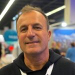 Person_25_Harald-Schmidt - University of Labour Mann mit kurzem, braunem Haar und lächelndem Gesicht, trägt einen schwarzen Hoodie, im Hintergrund unscharfe Messeatmosp...