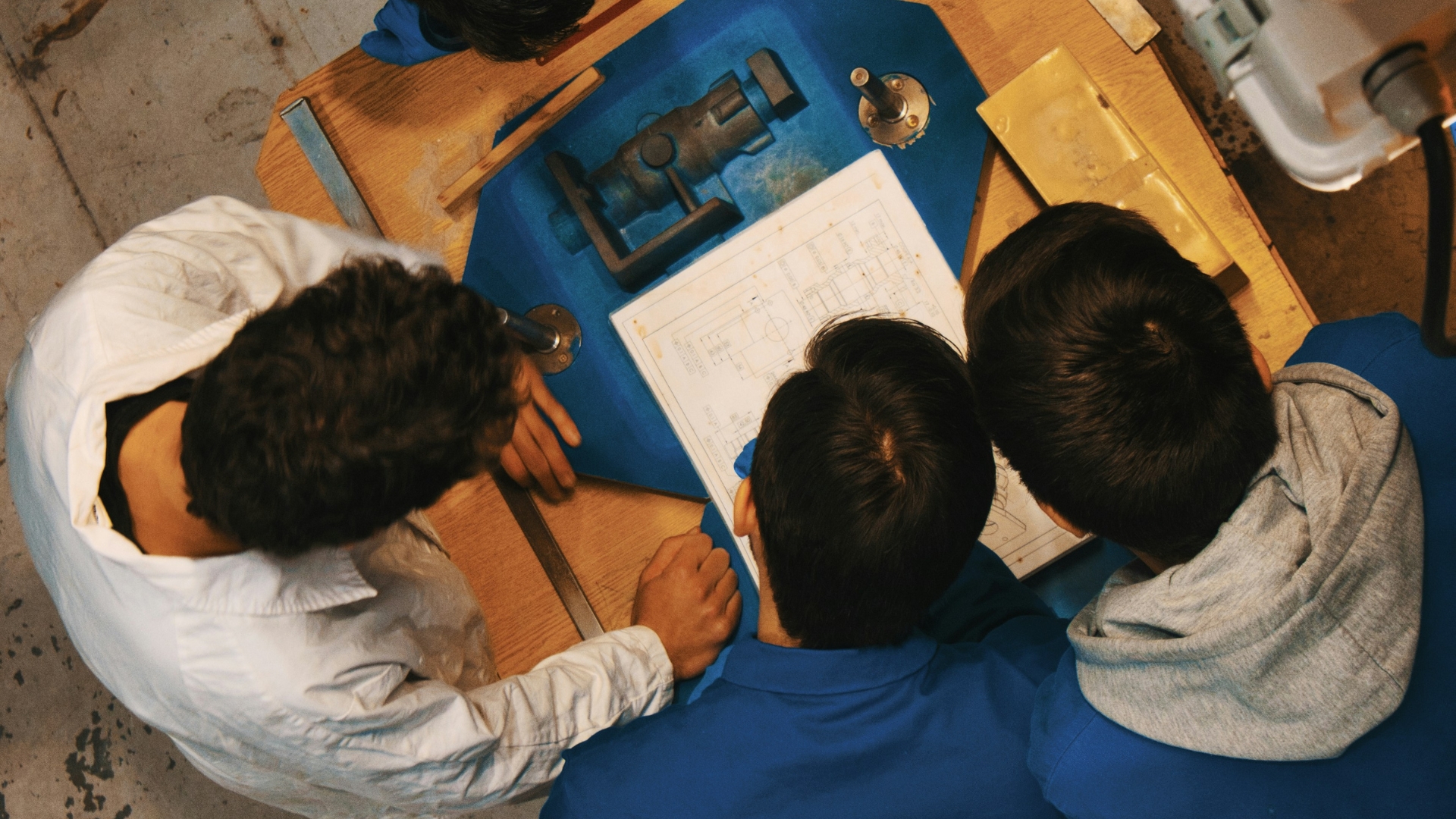 EV_25_Duale-Berufsbildung-Costa-Rica - University of Labour Drei Personen in Arbeitsanzügen betrachten gemeinsam technische Pläne auf einem Tisch. Das Bild ist von oben aufgenommen, ihre Köpfe sind nur von hinten zu sehen.