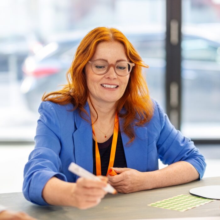 Frau mit rotem Haar und Brille in blauer Jacke, die an einem Tisch sitzt und mit einem Marker arbeitet.