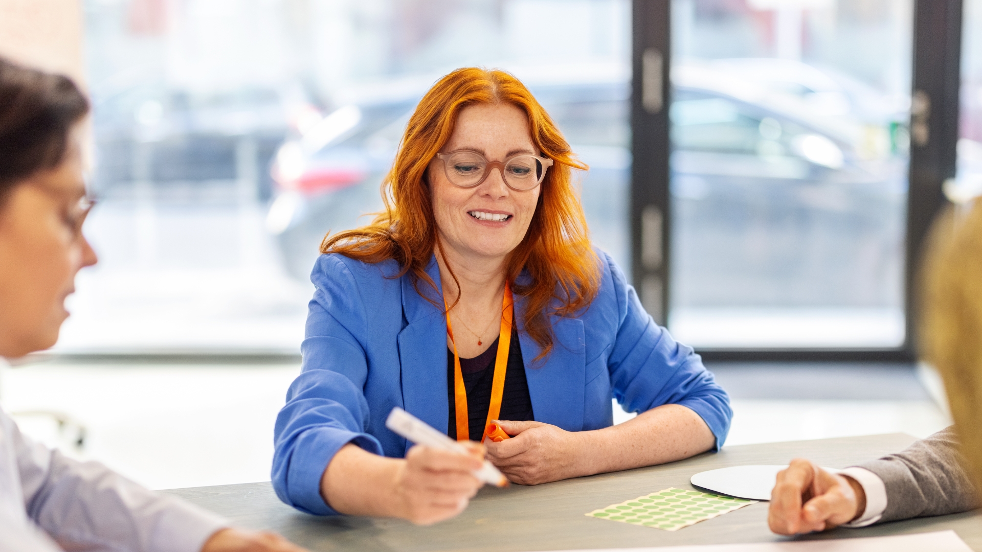 Frau mit rotem Haar und Brille in blauer Jacke, die an einem Tisch sitzt und mit einem Marker arbeitet.