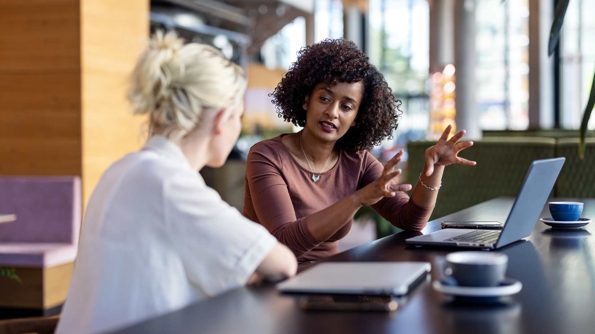 Zwei Frauen im Gespräch an einem Tisch, eine erklärt etwas gestikulierend.