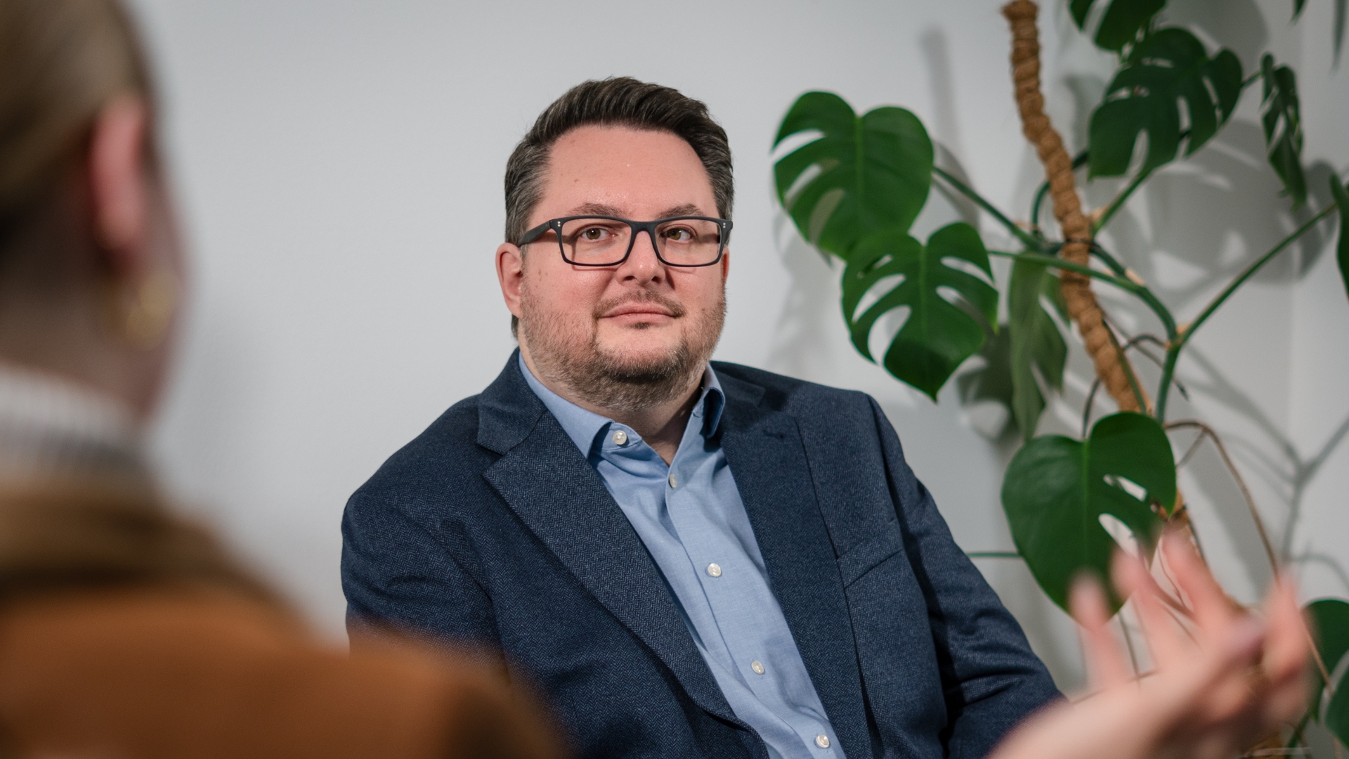 Heiko Hoßfeld sitzt in einer Gesprächssituation in der University of Labour. Im Hintergrund ist eine Pflanze zu sehen, im Vordergrund links unscharf eine Gesprächspartnerin.