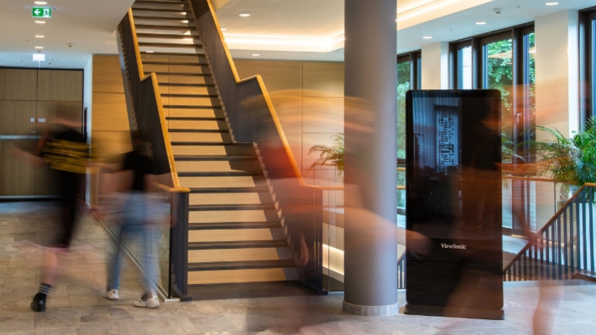 Bewegte Personen gehen eine Treppe in einem modernen Gebäude hinauf, umgeben von Pflanzen und großen Fenstern. Es handelt sich um das Foyer der University of Labour.