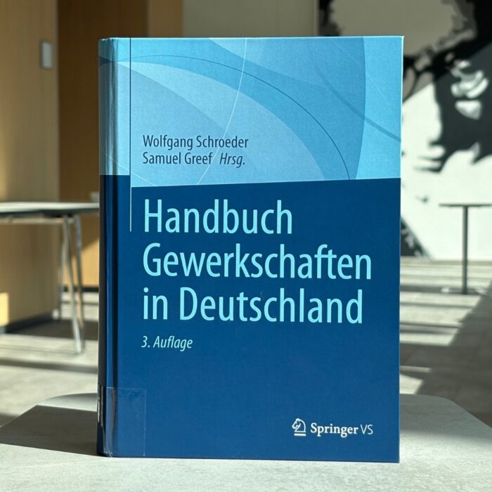 Auf einem Tisch im Foyer der University of Labour steht das Buch "Handbuch der Gewerkschaften". Im Hintergrund ist ein Gang sowie ein Wandbild von Hugo Sinzheimer zu erkennen.