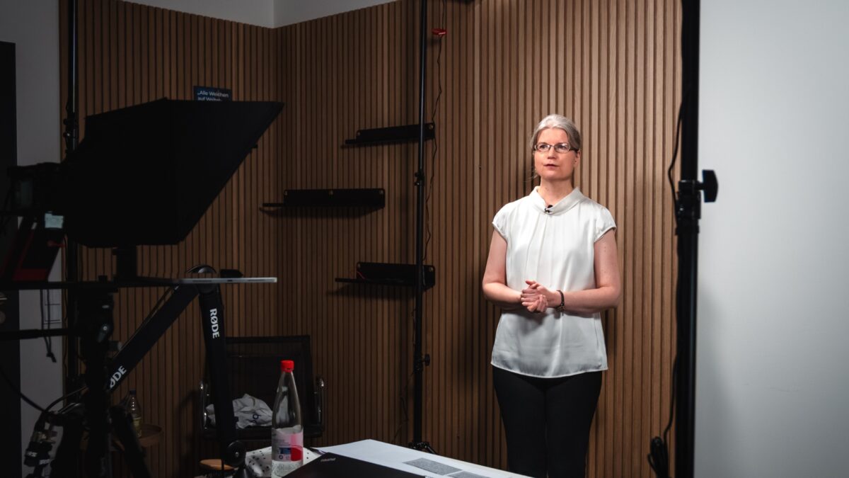 Ramona steht in einem Studio mit Holzpaneelen im Hintergrund. Links im Bild sind Stative und Kameras zu erkennen.