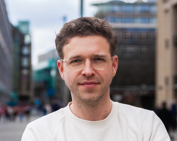 Portrait of a man with short brown hair and glasses, wearing a white shirt, standing on a city street.