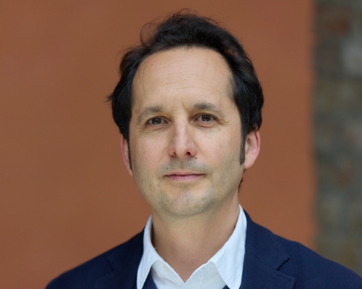Professional head-and-shoulders portrait of a man in a navy blazer and white shirt, neutral expression, orange blurred background