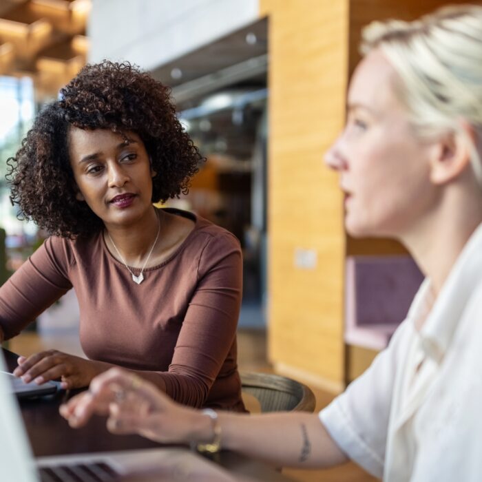 Zwei Frauen sitzen an einem Tisch mit Laptops, eine hört aufmerksam zu, während die andere spricht.