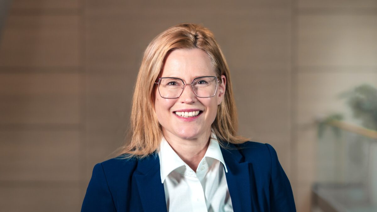 Wir sehen eine lächelnde Frau mit blonden Haaren und Brille in einem blauen Blazer. Im Hintergrund ist die Holzwand des Foyers der University of Labour zu erkennen.