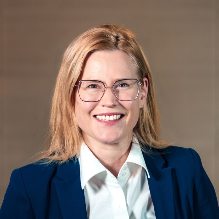 Wir sehen eine lächelnde Frau mit blonden Haaren und Brille in einem blauen Blazer. Im Hintergrund ist die Holzwand des Foyers der University of Labour zu erkennen.